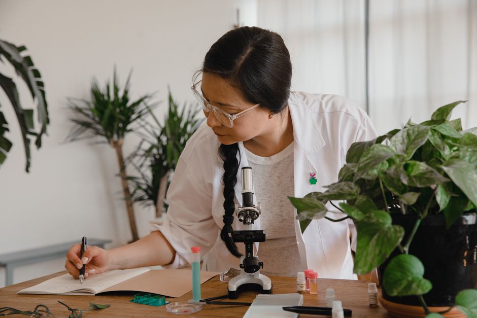 Female scientist conducting research and taking notes with microscope and plants.