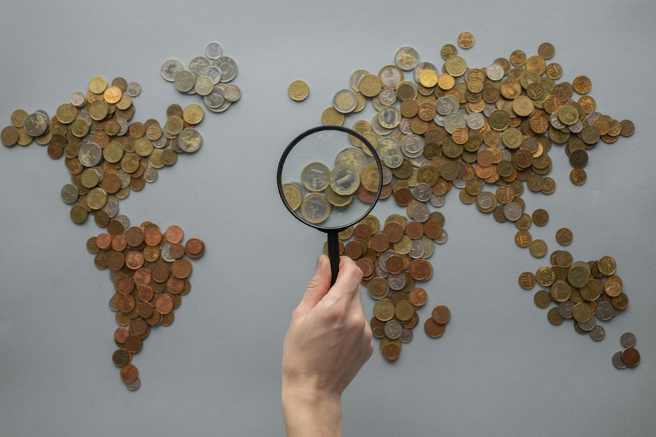 Top view of crop unrecognizable traveler looking through magnifying glass on world map made of various coins on gray background